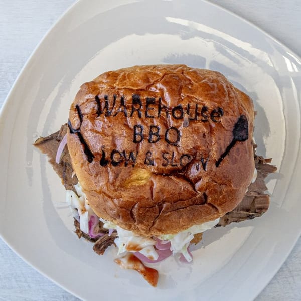 Warehouse BBQ's branded bun with pulled pork, coleslaw, and red onions on a white plate.