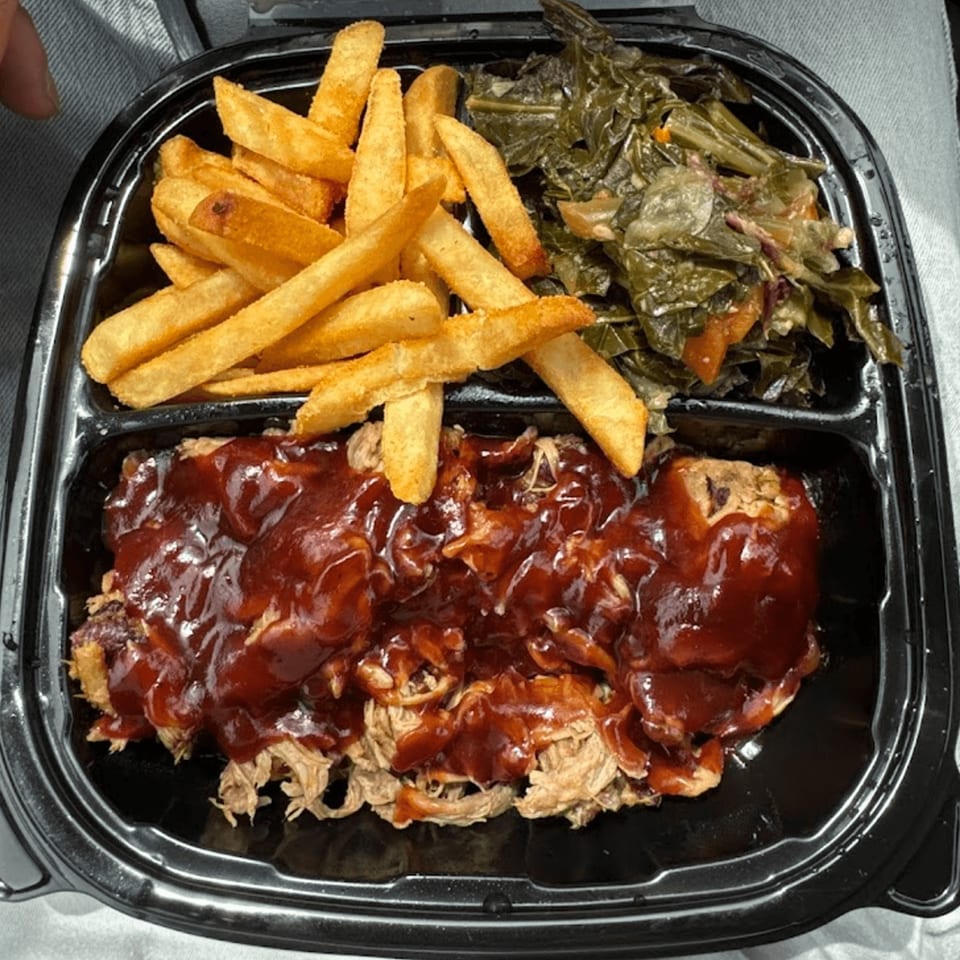 A takeout container with pulled pork covered in BBQ sauce, french fries, and collard greens.