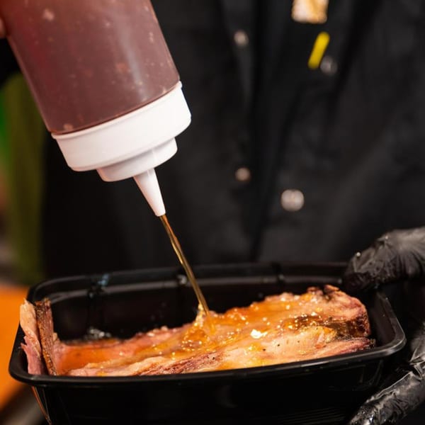A person in black gloves squeezes BBQ sauce from a bottle onto a slab of meat.