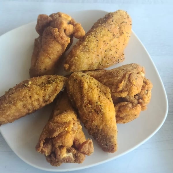 A white plate holds several golden-brown, crispy chicken wings, seasoned with black pepper.
