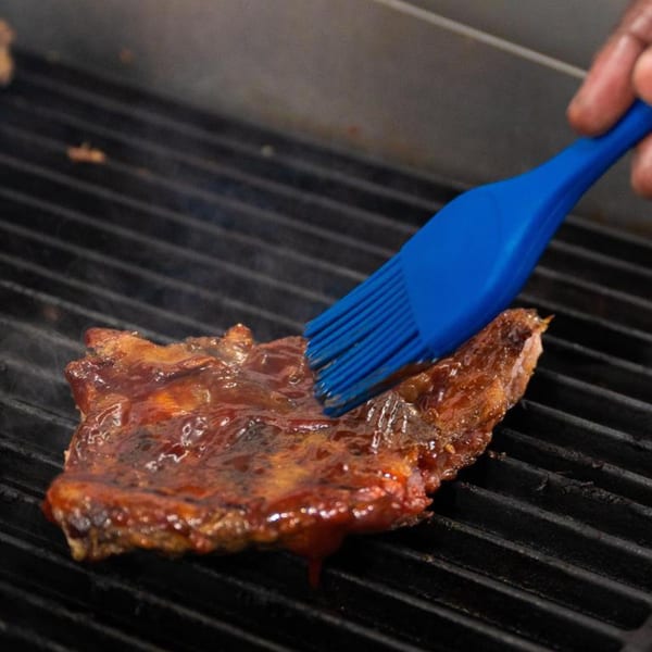 A hand brushes barbecue sauce onto a rack of ribs cooking on a grill.